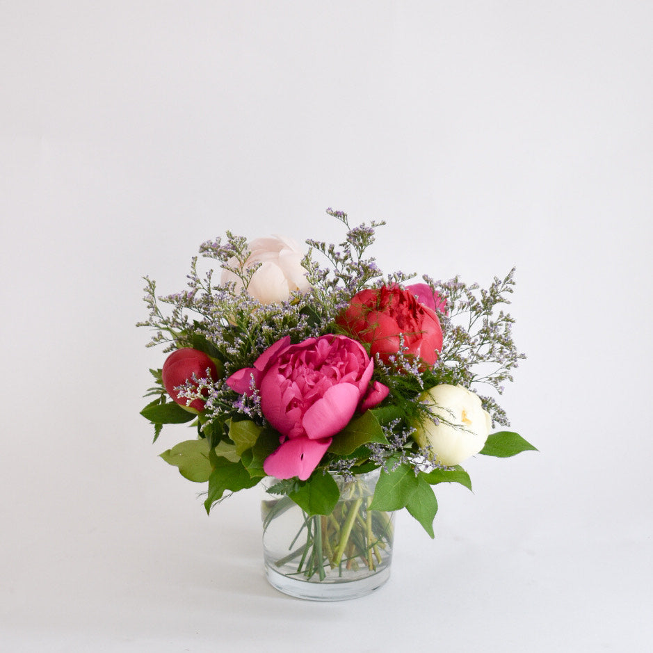 A medium-stemmed vase arrangement of peony blooms in various colors including pink and white, with green leaves and seasonal fillers.