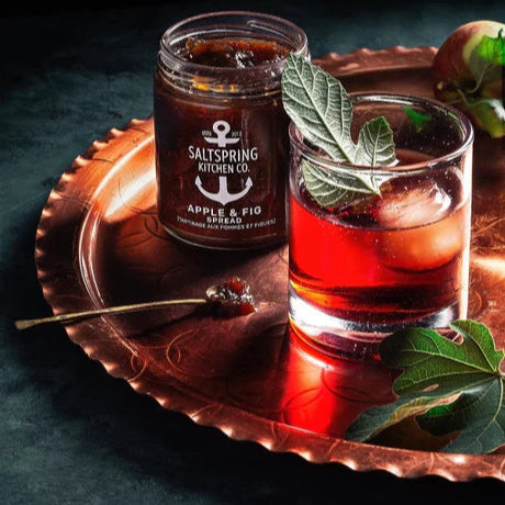 Jar of 'Apple & Fig' preserve next to a glass of red drink with ice and mint on a copper tray.