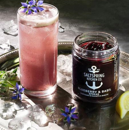 Pink cocktail with a blue flower garnish next to a jar of Saltspring Kitchen Co. blueberry and basil spread on a reflective surface.