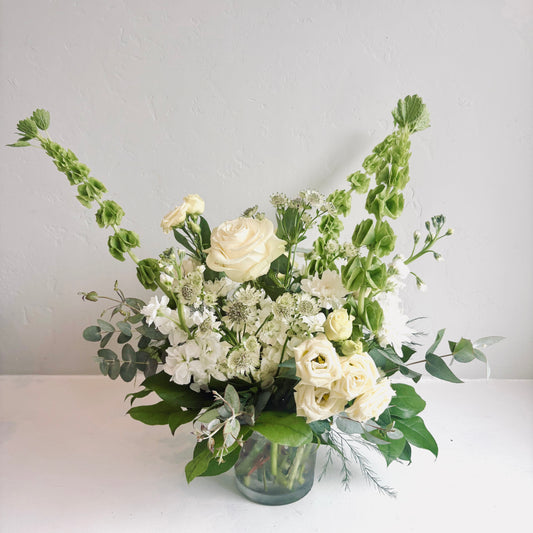 A floral arrangement in a clear glass vase, featuring cream-colored roses, white filler flowers, green foliage, and tall green stems, set against a plain light gray wall and white surface.