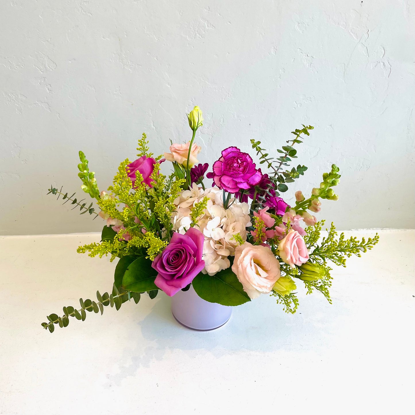 A small pink vase arrangement using assorted seasonal  flowers in shades of white, pink, cream, lavender and purples. Designed in a whimsical, garden style. 