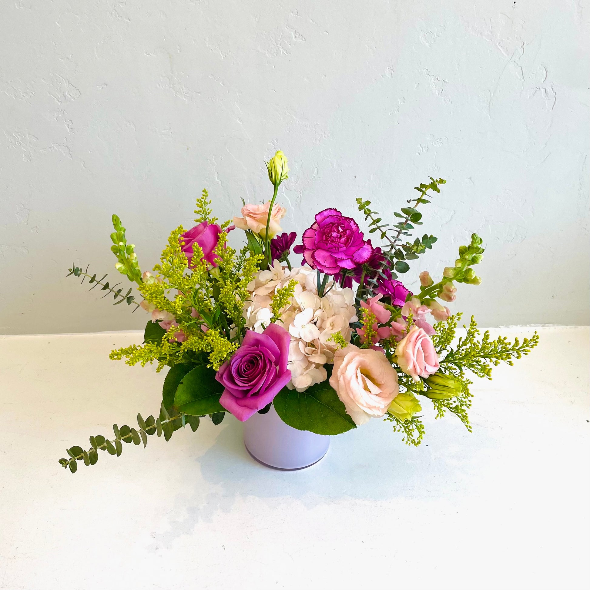 A small pink vase arrangement using assorted seasonal  flowers in shades of white, pink, cream, lavender and purples. Designed in a whimsical, garden style. 