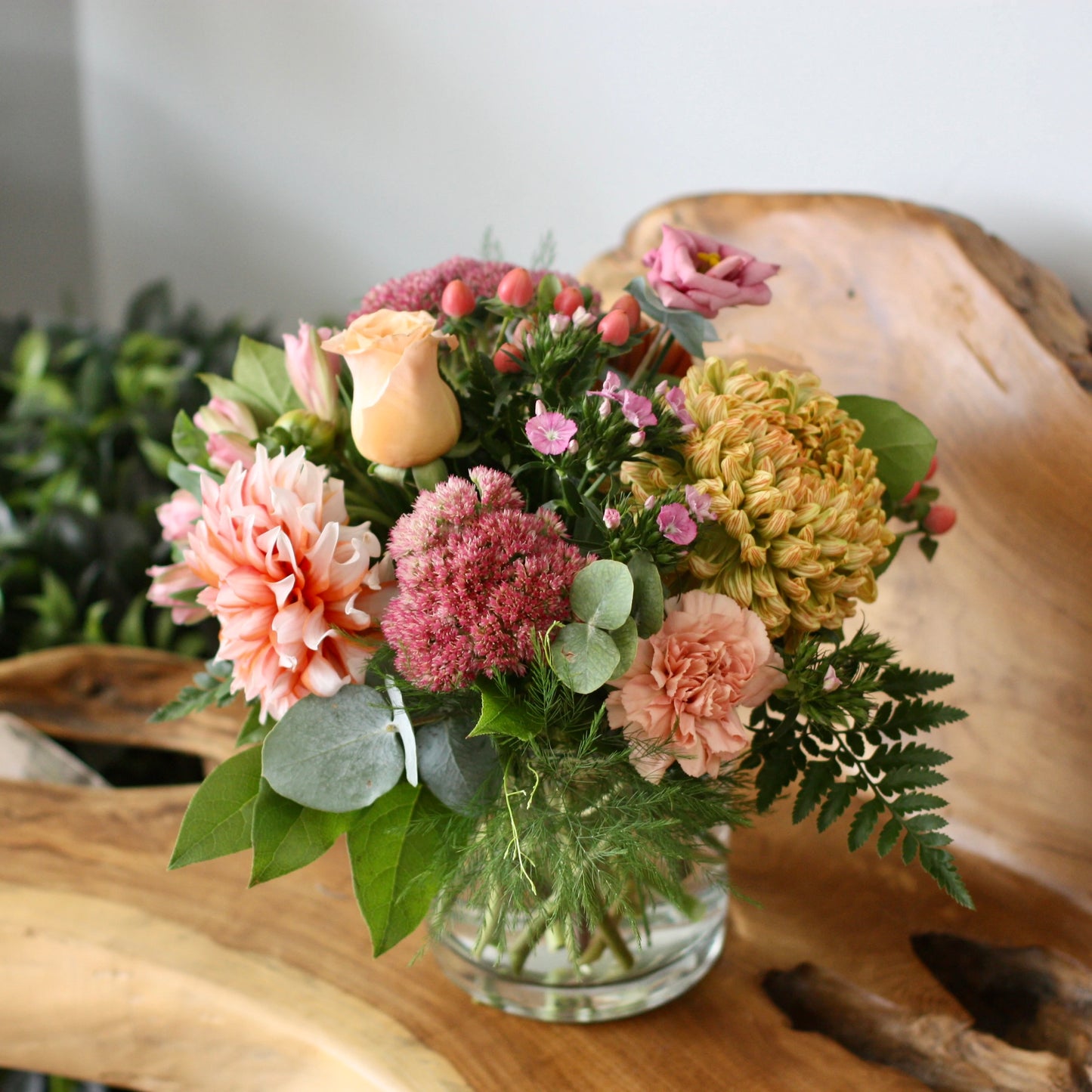 A floral arrangement featuring a variety of flowers in seasonal colors, presented in a glass vase, with greenery and a lush background.